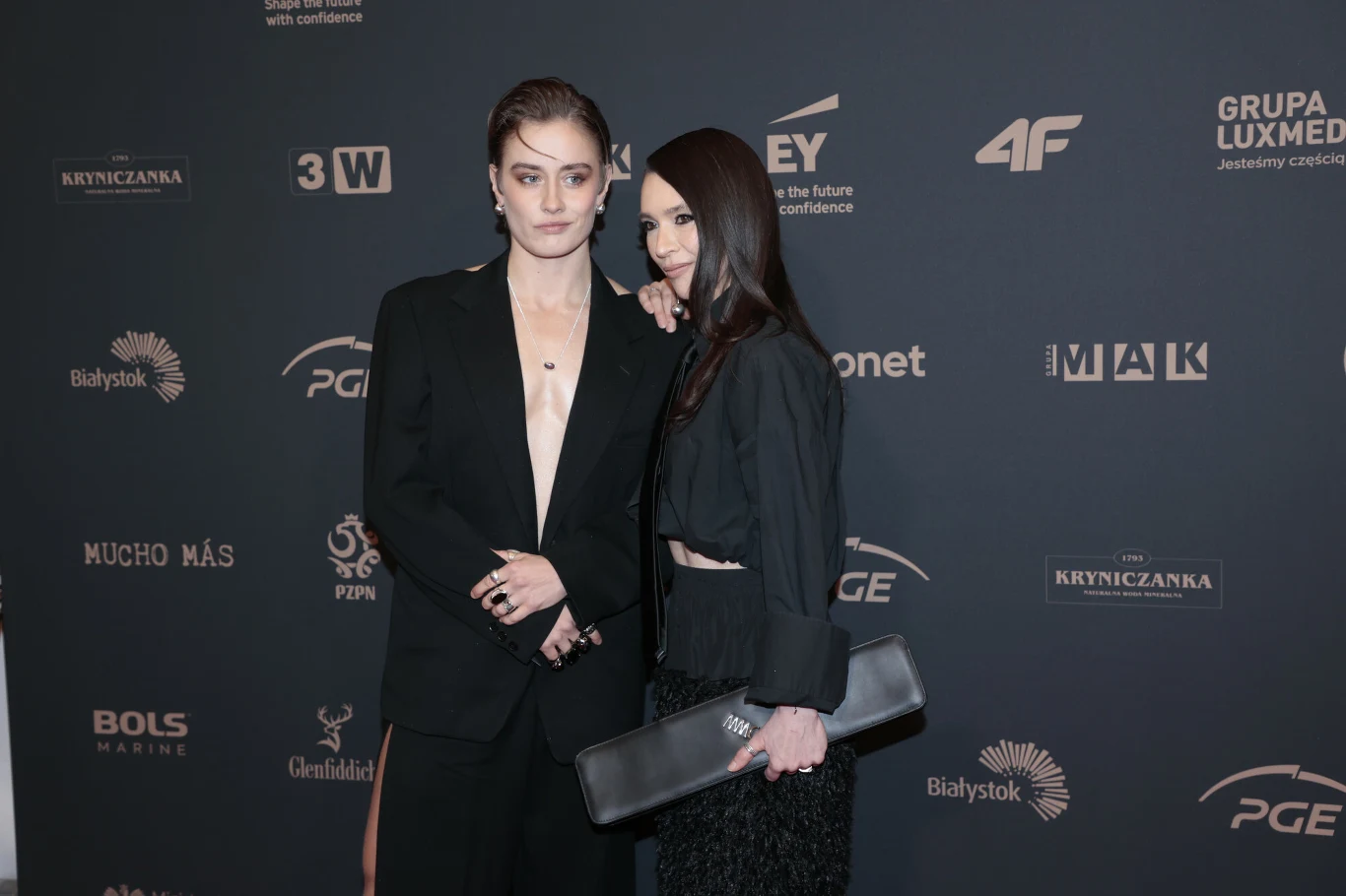 Dwie elegancko ubrane kobiety pozują razem na ściance z ciemnym tłem z widocznymi logotypami sponsorów i partnerów wydarzenia.