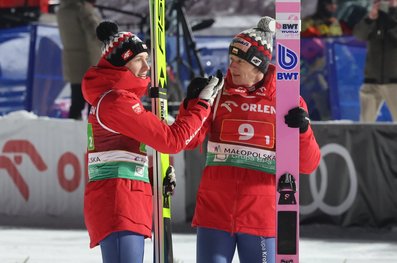 Dawid Kubacki i Kacper Tomasiak (po lewej) Dwóch skoczków narciarskich ubranych w czerwone kombinezony i czapki zimowe rozmawia ze sobą na skoczni. Jeden z nich trzyma narty, w tle widać elementy infrastruktury sportowej oraz innych uczestników zawodów.