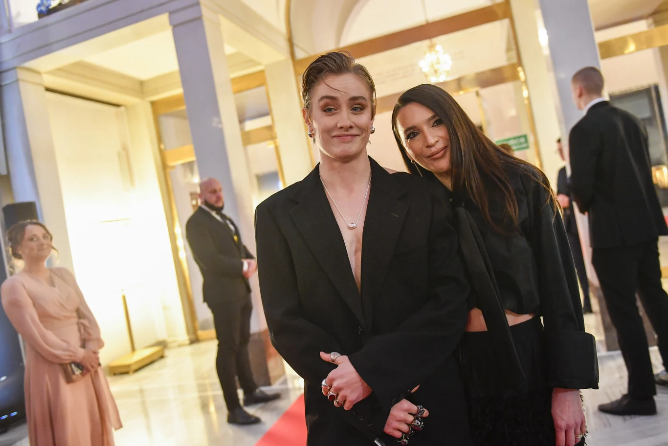 Dwie elegancko ubrane osoby stoją blisko siebie na czerwonym dywanie w jasnym, ozdobnym wnętrzu, w tle widoczni są inni uczestnicy wydarzenia oraz ochrona.