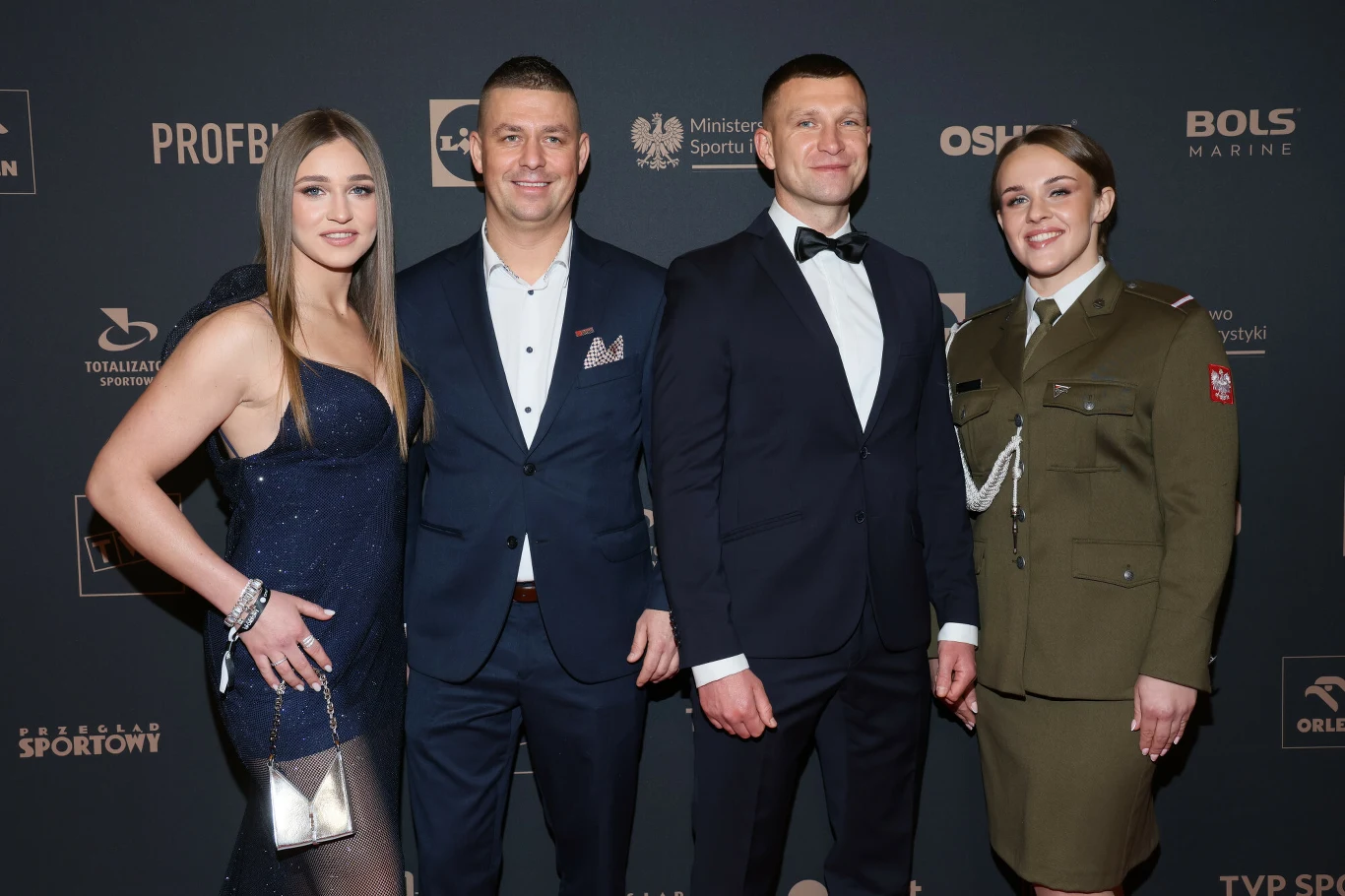 Czworo elegancko ubranych osób pozuje na tle ściany z logotypami sponsorów i organizatorów. Dwie kobiety stoją po bokach, jedna w sukience wieczorowej, druga w mundurze, a dwóch mężczyzn w garniturach znajduje się między nimi.