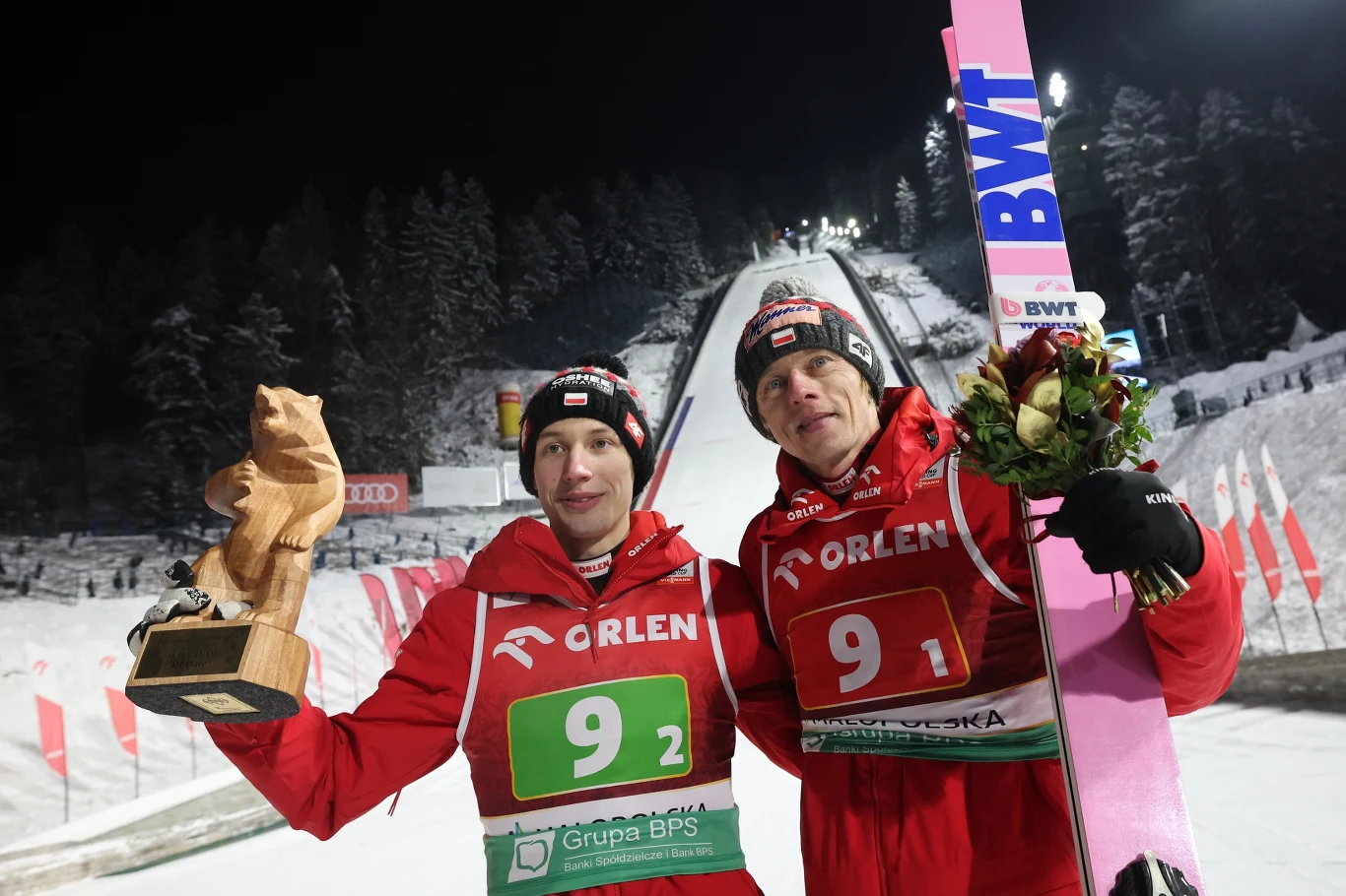 Kacper Tomasiak (po lewej) i Dawid Kubacki ze swoimi nagrodami Dwóch skoczków narciarskich w czerwonych strojach stoi na tle skoczni narciarskiej, jeden trzyma puchar w kształcie zwierzęcia, drugi wiązankę kwiatów i narty, wokół śnieg i zimowy krajobraz.