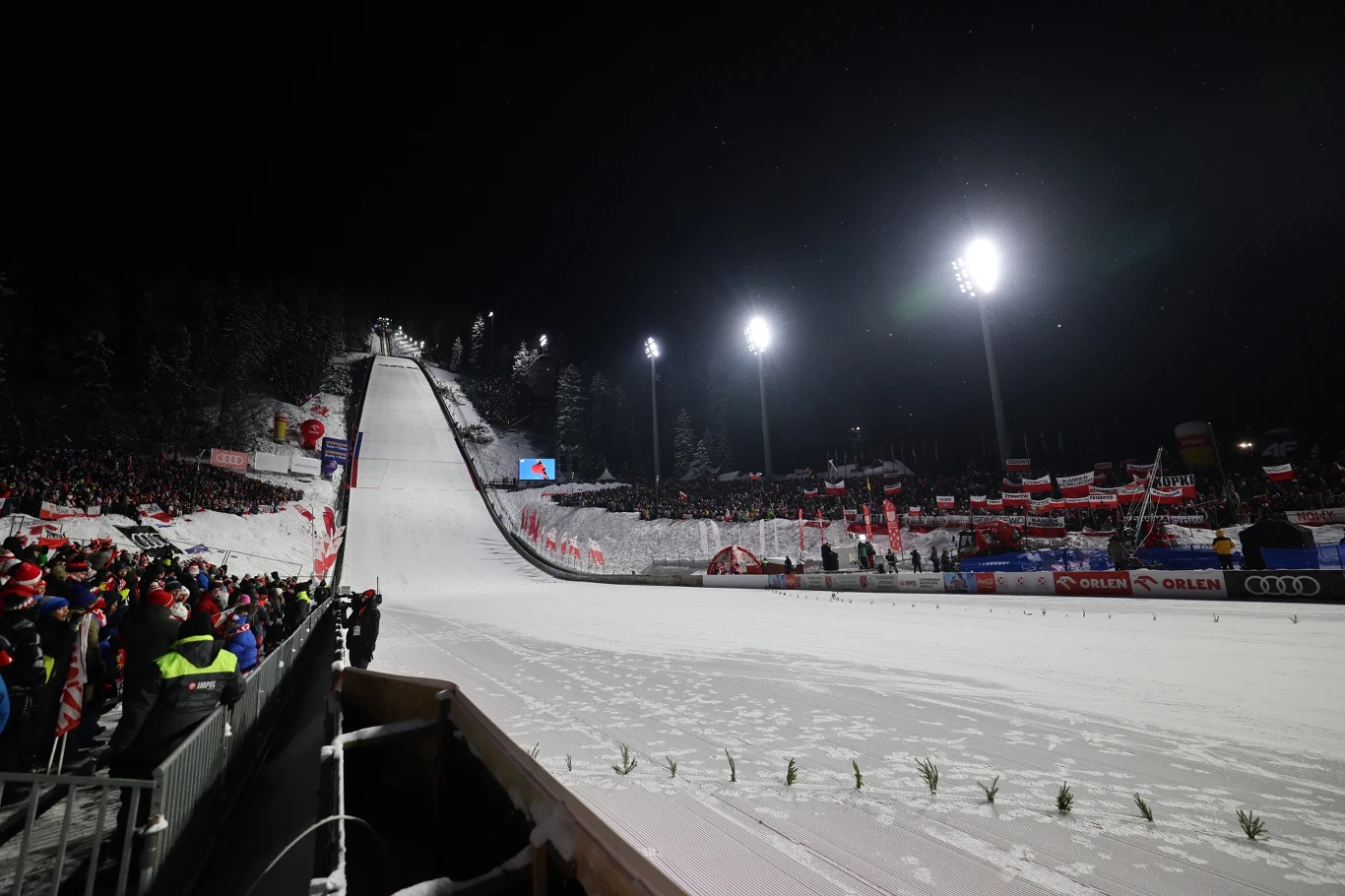 Oświetlona nocą skocznia narciarska otoczona tłumami kibiców z flagami i trybunami, zaśnieżony teren przygotowany do zawodów skoków narciarskich, atmosfera sportowego święta.