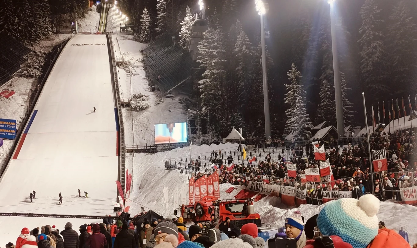 Tłum kibiców zgromadzony wokół skoczni narciarskiej w otoczeniu ośnieżonych drzew, jasne oświetlenie stadionowe, flaga Polski i inne flagi państwowe wśród publiczności, na skoczni widoczni sportowcy, atmosfera sportowego wydarzenia zimowego.