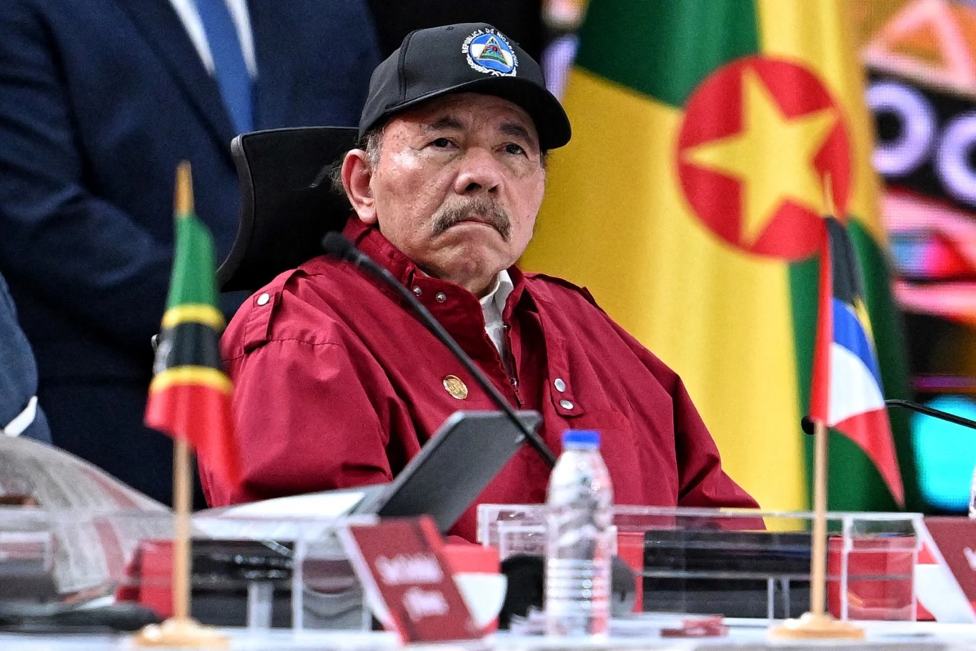 Nikaragua uwolniła kilkadziesiąt osób. MSW wydało komunikat Starszy mężczyzna w czerwonej kurtce i czapce siedzi za stołem konferencyjnym, wokół rozmieszczone są flagi państwowe oraz mikrofony, w tle widoczne kolorowe flagi.