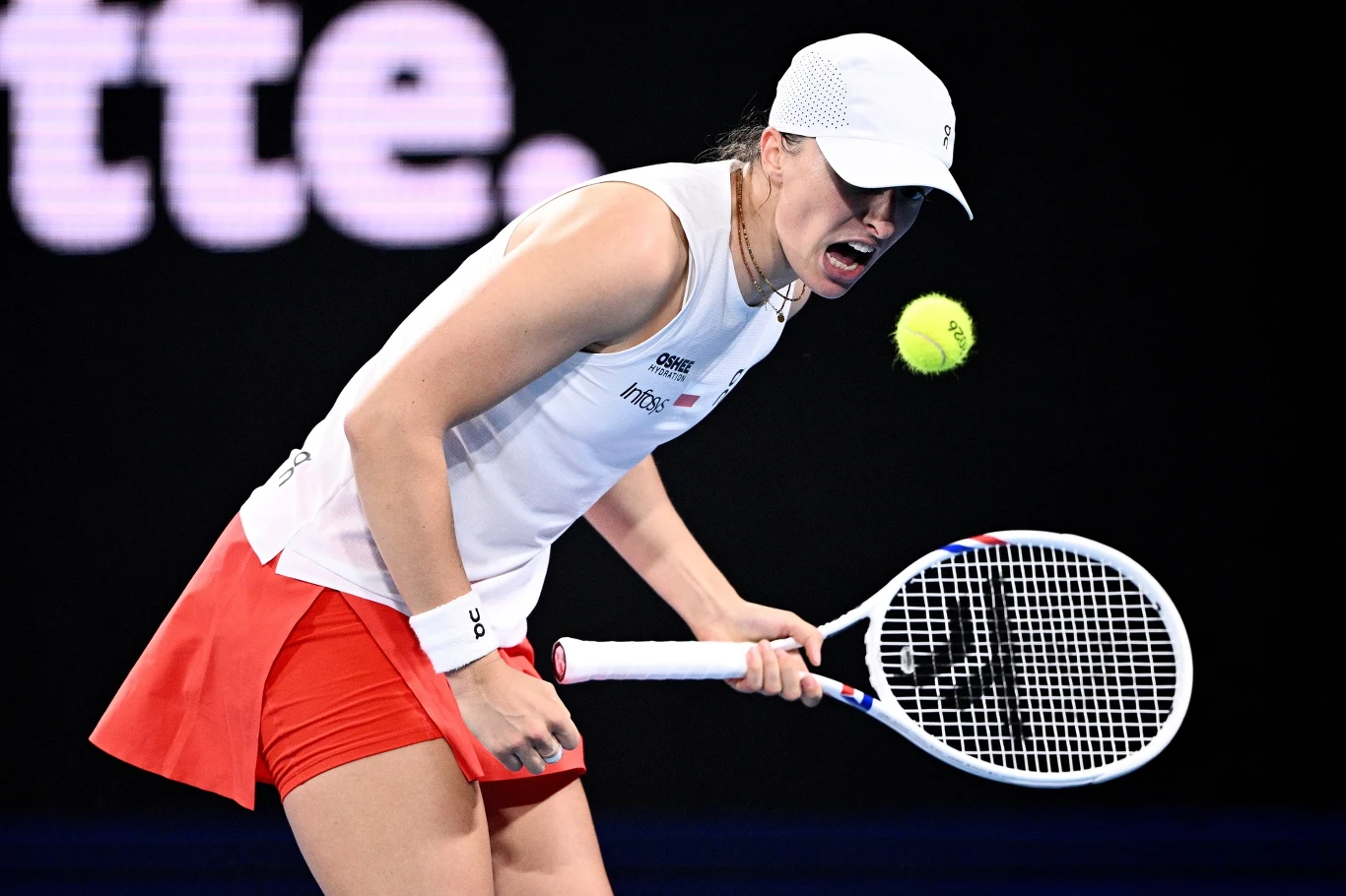 Iga Świątek w meczu z Belindą Bencic Tenisistka uderzająca piłkę podczas meczu na korcie, ubrana w białą czapkę, białą koszulkę i czerwoną spódnicę, skupiona na akcji.