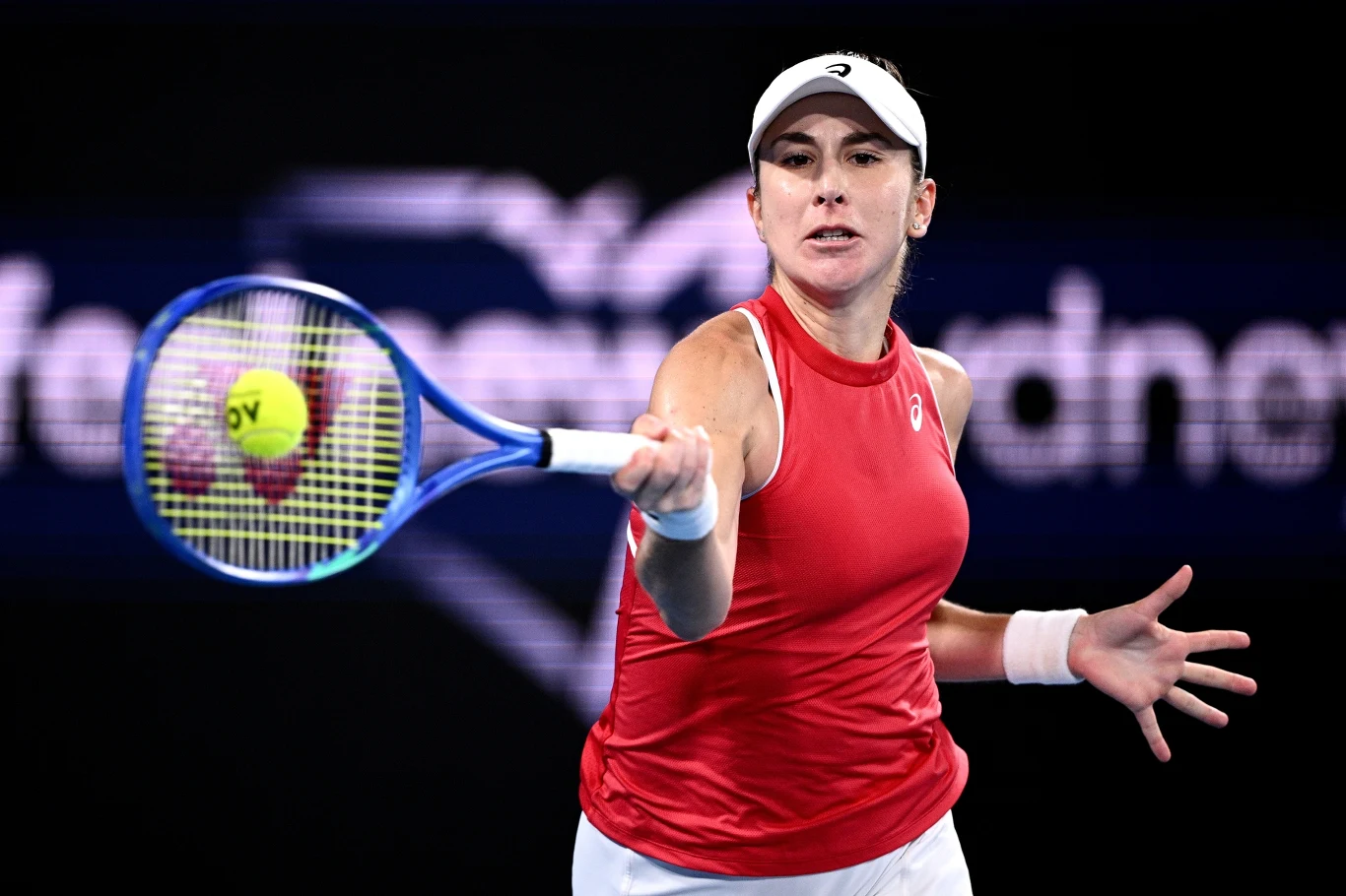 Belinda Bencic Tenisistka w czerwonym stroju i białej czapce odbija piłkę rakietą podczas meczu na korcie, w tle rozmyte logo sponsora.