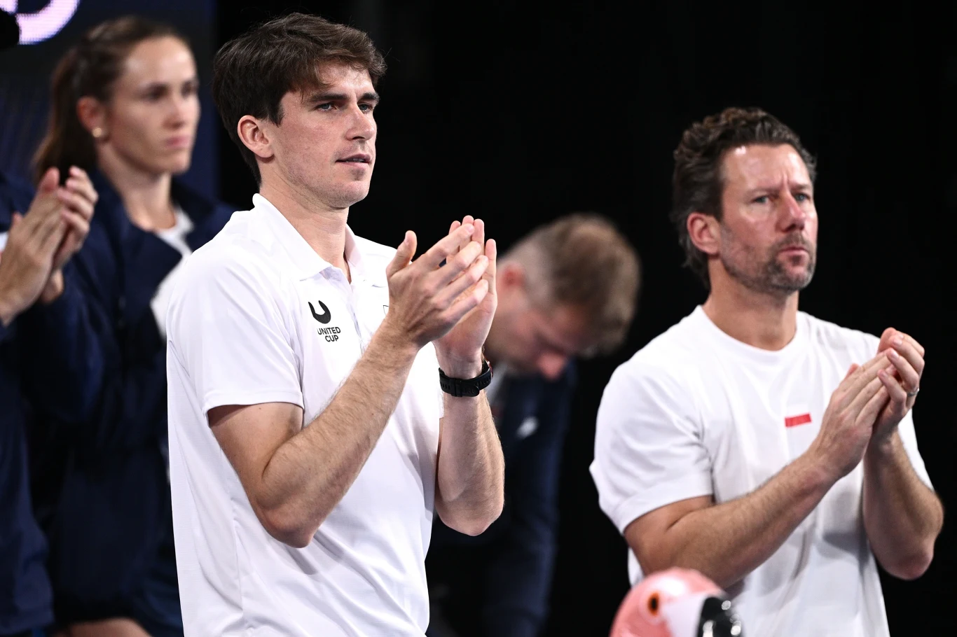 Kapitan Mateusz Terczyński i zaniepokojony Wim Fissette w polskim boksie Dwie osoby w białych koszulkach stoją obok siebie i klaszczą, w tle widoczne są inne postacie, część z nich niewyraźnie. Atmosfera sportowego wydarzenia, skupienie i zaangażowanie na twarzach.