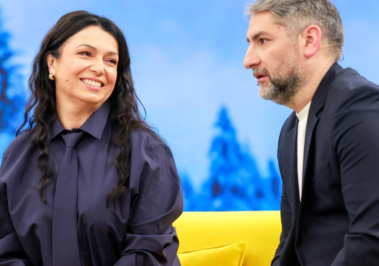 Katarzyna Pakosińska, Irakli Basilaszwili Dwie osoby siedzą na żółtej kanapie obserwując się nawzajem, kobieta uśmiecha się szeroko, mężczyzna patrzy poważnie, w tle widoczna niebieska sceneria z zarysem drzew.