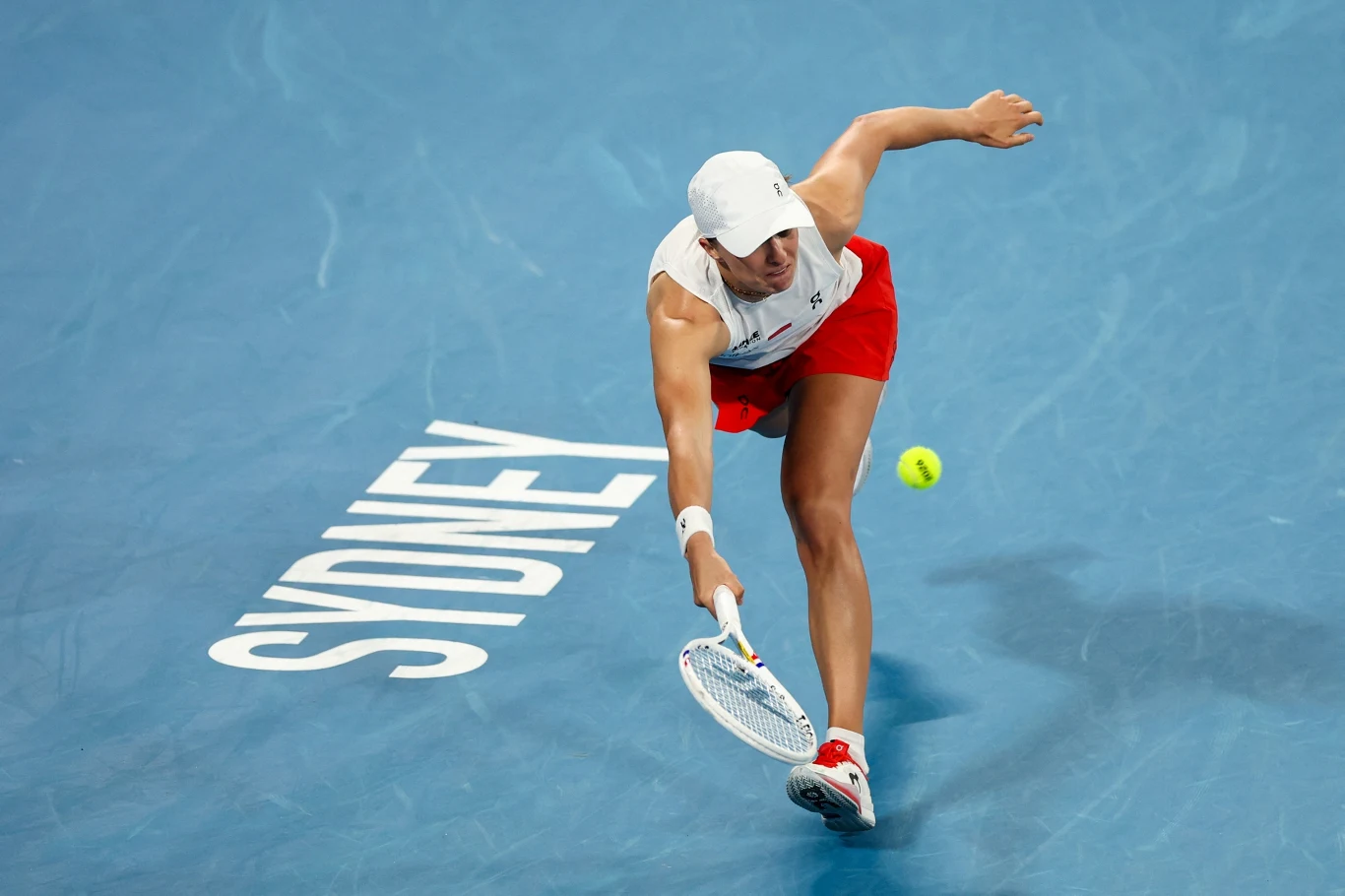 Tenisistka w krótkich czerwonych spodenkach i białej koszulce podczas dynamicznego uderzenia piłki tenisowej na niebieskim korcie z napisem SYDNEY.