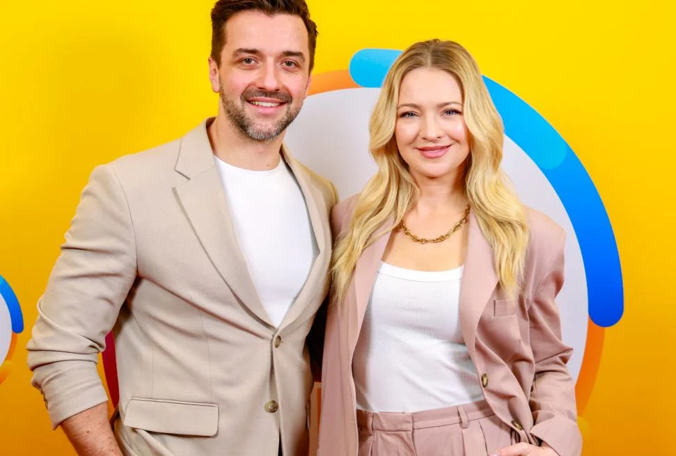 Rafał Szatan i Barbara Kurdej-Szatan Dwoje uśmiechniętych dorosłych ubranych w eleganckie, jasne garnitury stoi na tle żółtej ścianki z kolorowymi elementami graficznymi.