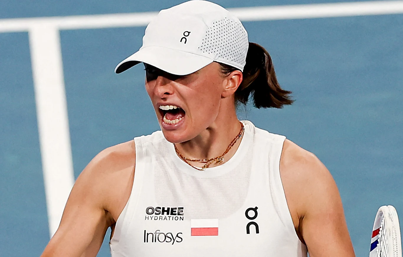 Iga Świątek Zawodniczka tenisowa w białej koszulce i czapce sportowej wyraża silne emocje po zakończonej akcji na korcie tenisowym. W tle jasnoniebieska nawierzchnia kortu, widoczny fragment rakiety.