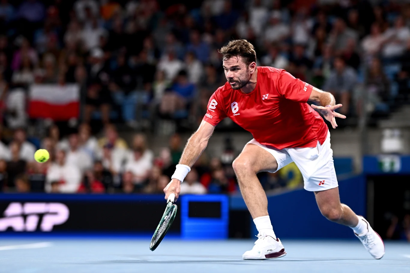 Zawodnik tenisowy w czerwonej koszulce pochyla się, próbując odbić piłkę rakietą tuż nad kortem. W tle widoczna publiczność oraz polska flaga.