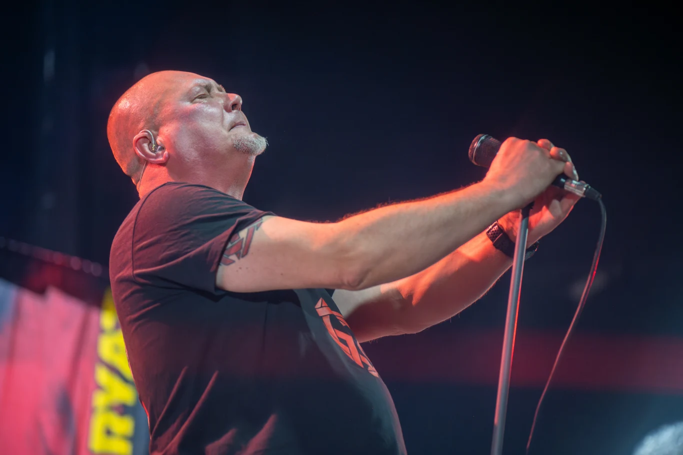 Tomasz "Oley" Olejnik stoi na czele grupy Proletaryat Mężczyzna łysy śpiewa do mikrofonu na scenie koncertowej, ma tatuaż na ramieniu i nosi czarną koszulkę, tło ciemne z rozmytym banerem.