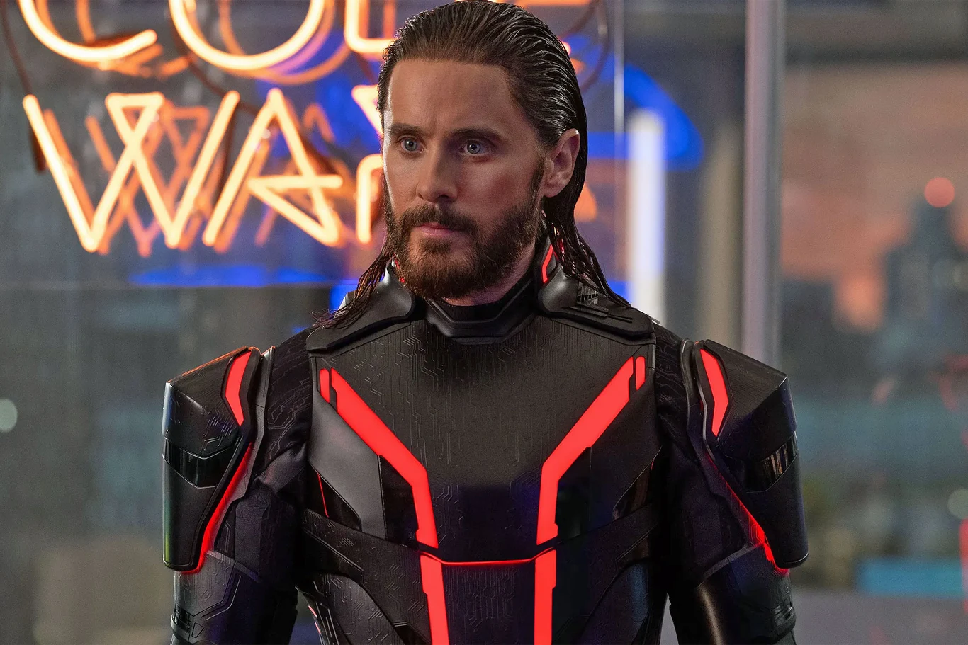 Mężczyzna z brodą i długimi włosami ubrany w futurystyczny czarny strój z czerwonymi akcentami, stojący we wnętrzu z neonowym napisem w tle.