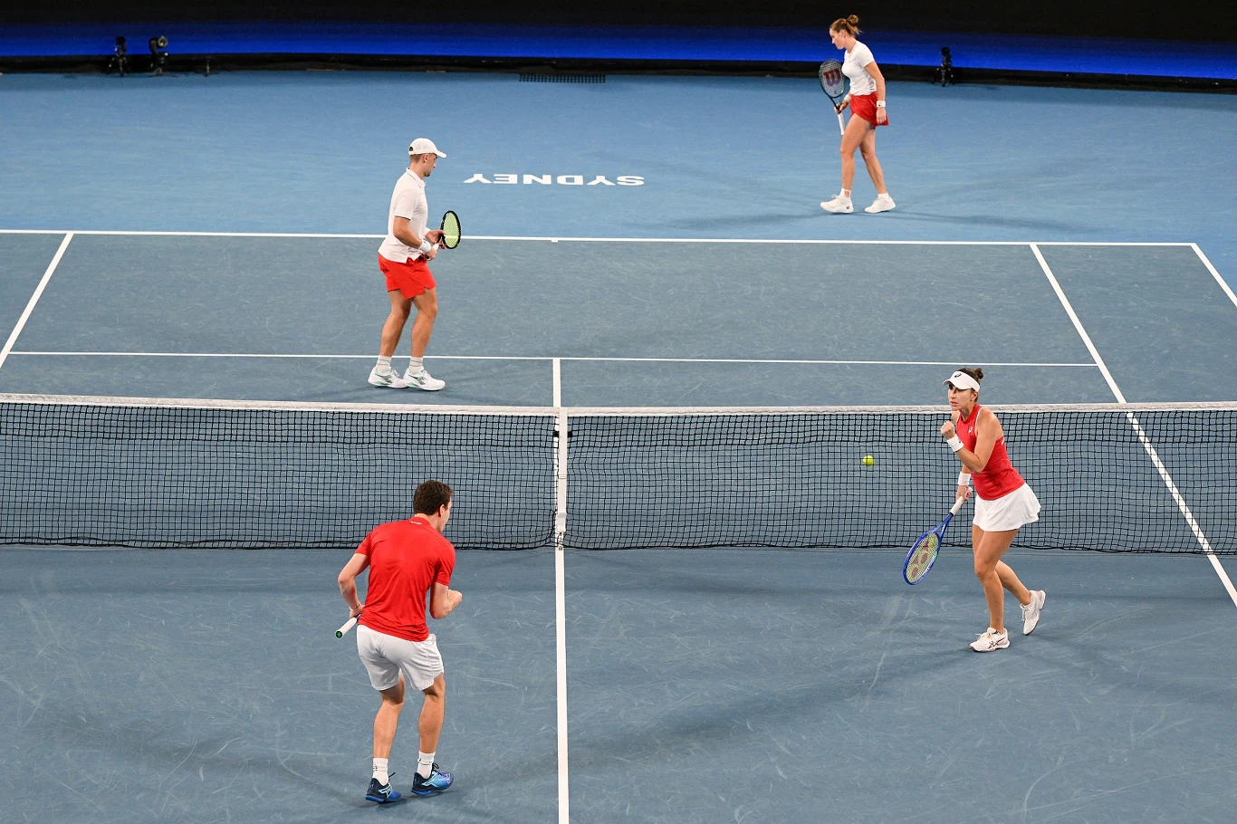 Czterech zawodników podczas meczu deblowego w tenisie na niebieskim korcie, ubrani w czerwone i białe stroje sportowe, piłka w locie, w tle widoczny napis 'Sydney'.