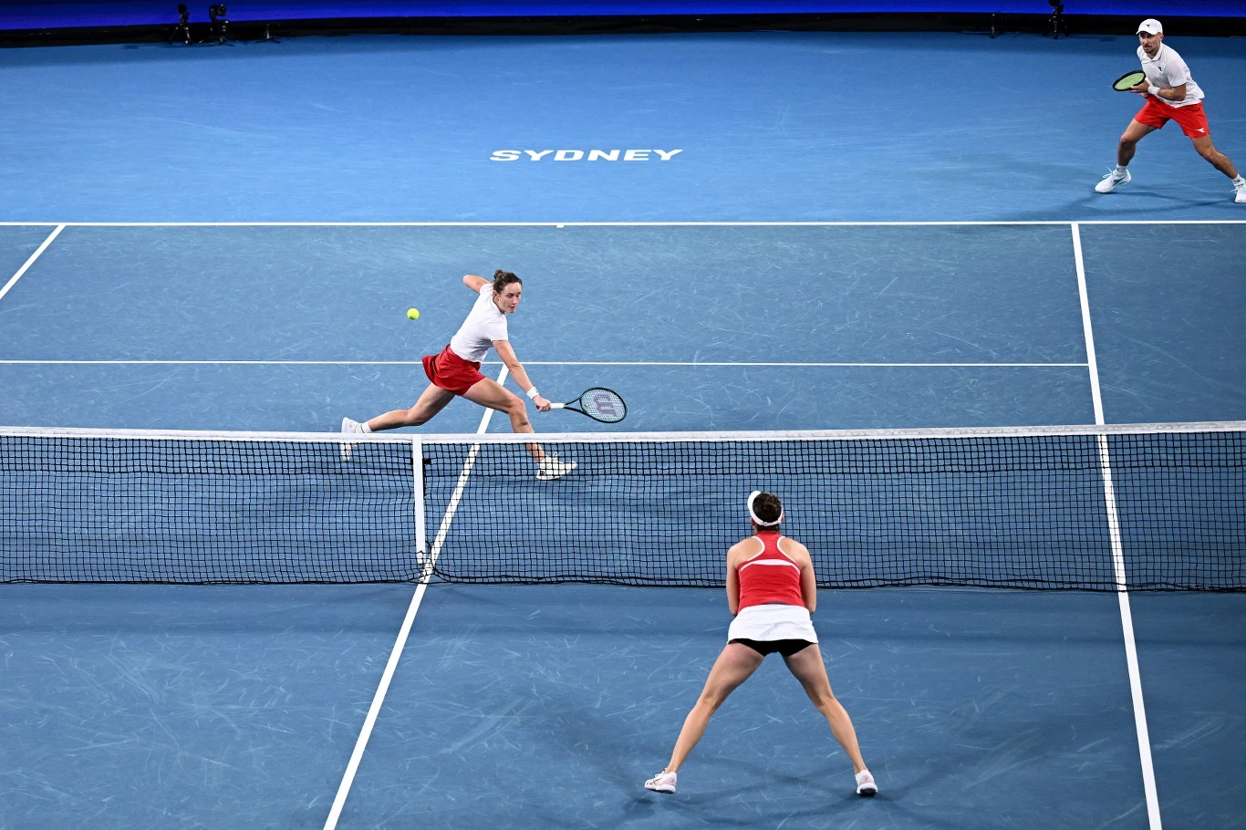 Trzech tenisistów w trakcie meczu deblowego na niebieskim korcie, dwóch z nich w białych koszulkach i czerwonych spodenkach po jednej stronie siatki, jedna zawodniczka w biało-czerwonym stroju, dynamiczna akcja z piłką w locie, na korcie napis Sydney.
