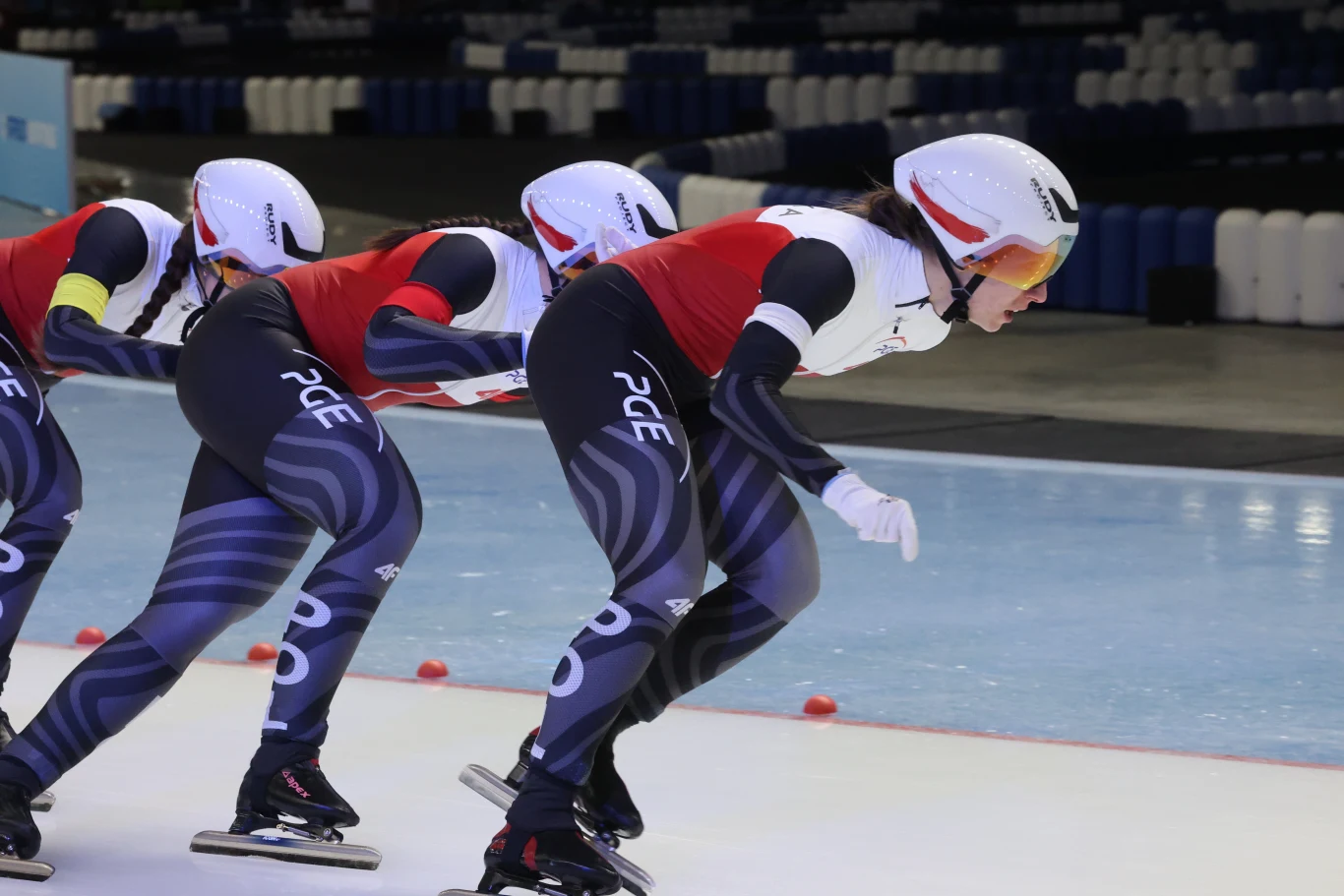 Zawodniczki drużyny panczenistów podczas jazdy szybkiej na lodzie, ubrane w stroje startowe, kaski i okulary ochronne, koncentrujące się na technice i utrzymaniu prędkości na lodowisku.
