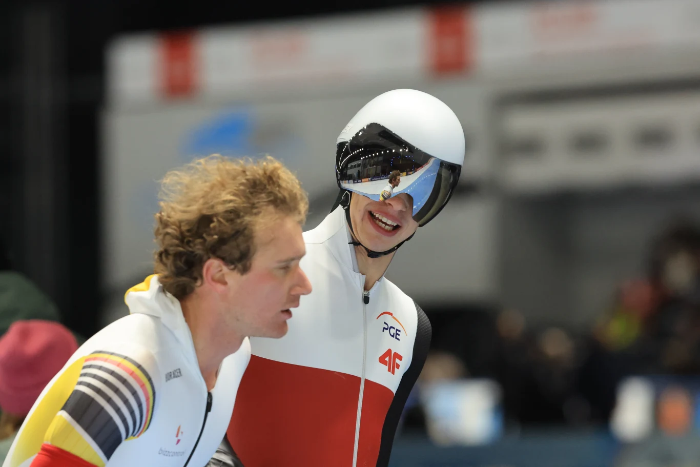 Dwóch łyżwiarzy wyścigowych w strojach reprezentacyjnych, jeden uśmiecha się, a drugi patrzy w bok, w tle rozmyte sylwetki innych osób i trybuny.