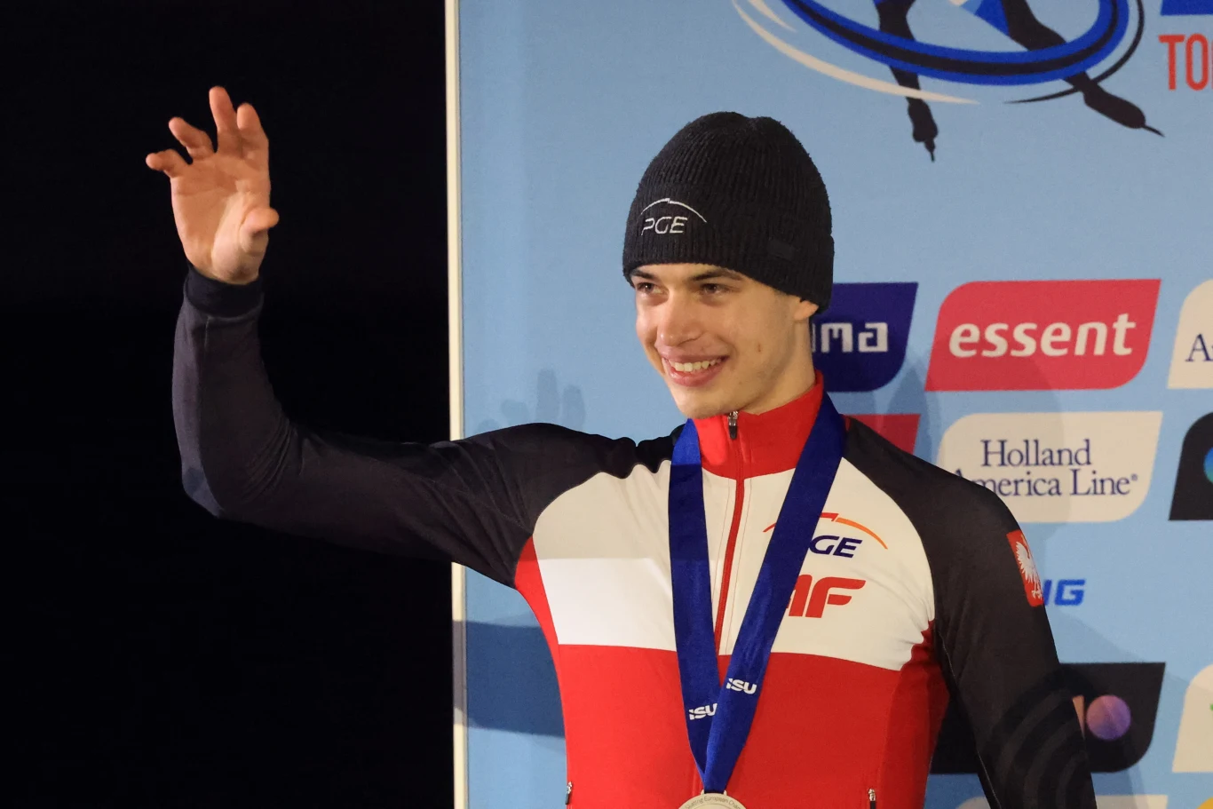 Sportowiec w czerwono-białym stroju oraz czarnej czapce, z medalem zawieszonym na szyi, uśmiecha się i unosi rękę w geście pozdrowienia. W tle plansza z logotypami sponsorów.