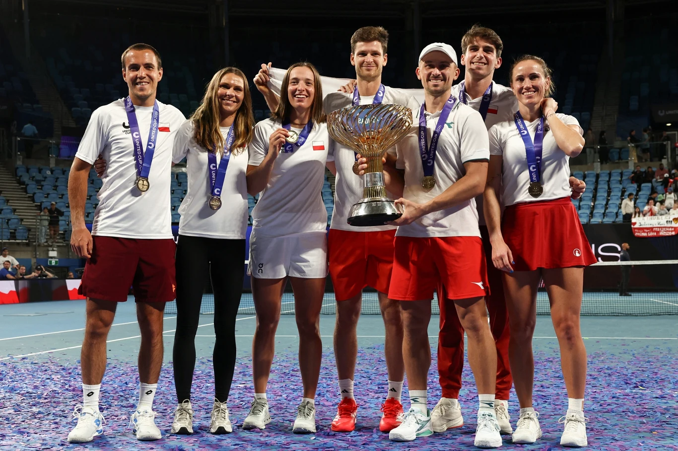 Polska triumfatorem United Cup AD 2026 Grupa siedmiu uśmiechniętych tenisistów w sportowych strojach z medalami na szyjach i dużym pucharem trzymanym centralnie, stojąca razem na korcie tenisowym po wygranym turnieju.