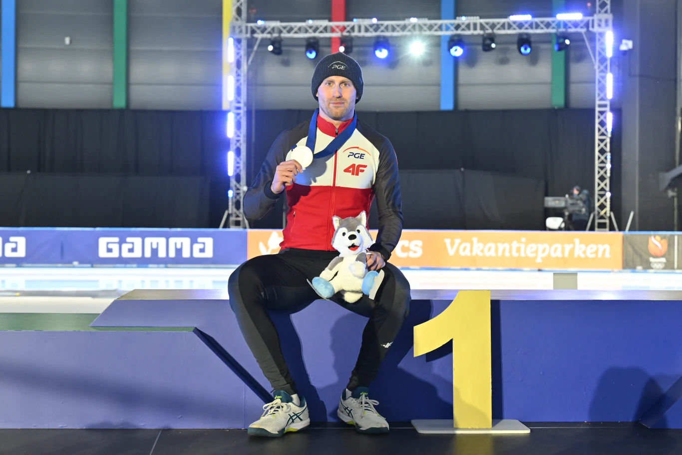 Sportowiec w stroju reprezentacyjnym siedzi na podium na pierwszym miejscu, trzyma medal oraz maskotkę, w tle elementy hali sportowej z oświetleniem.