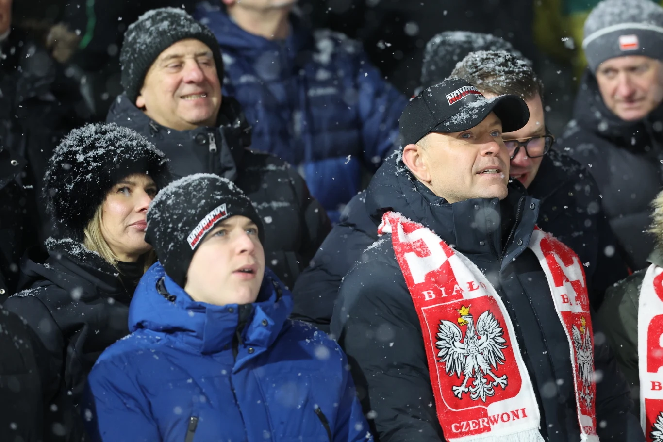 Grupa ludzi ubrana w zimowe ubrania podczas opadów śniegu na stadionie, kibice trzymający biało-czerwone szaliki z godłem Polski, atmosfera wsparcia dla drużyny sportowej.