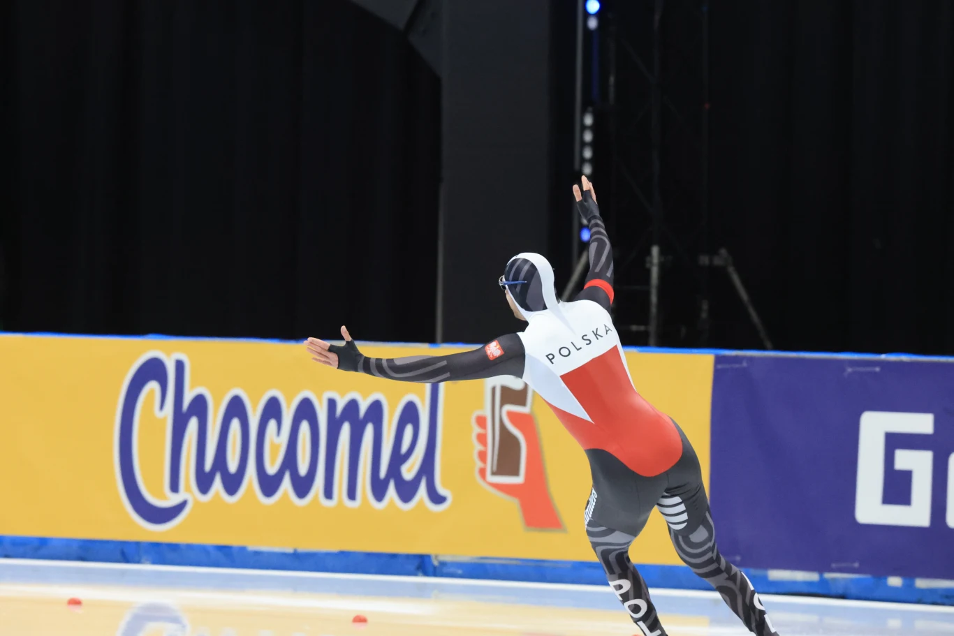 Łyżwiarz w stroju reprezentacji Polski dynamicznie sunie po lodowisku podczas zawodów, w tle widoczne są barwne reklamy na bandach oraz czarna kurtyna.