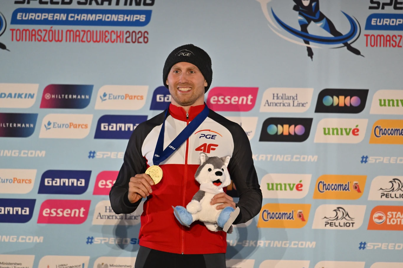 Mężczyzna w sportowym stroju z uśmiechem na twarzy trzyma złoty medal oraz maskotkę, stojąc na tle ściany z logotypami sponsorów i nazwą zawodów Mistrzostwa Europy w łyżwiarstwie szybkim w Tomaszowie Mazowieckim 2026.