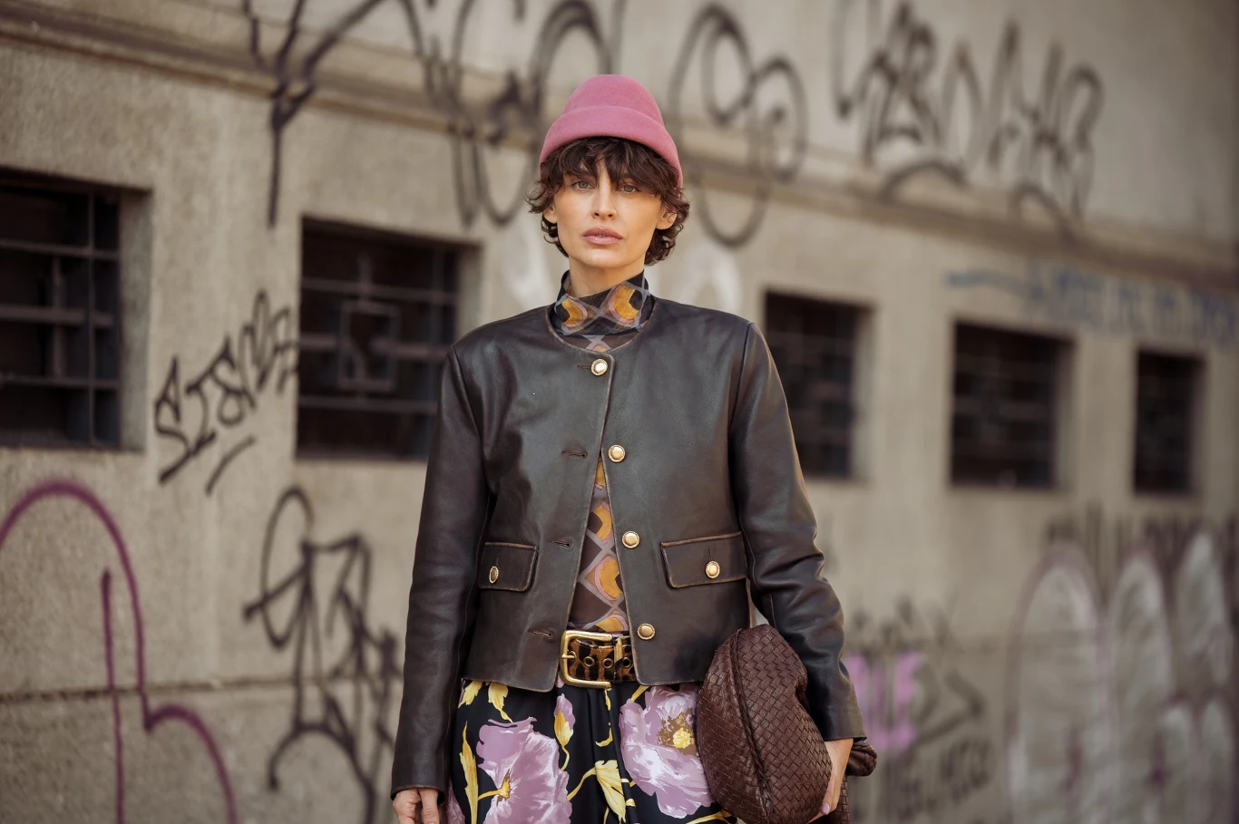 Trendy w modzie na 2026 rok Osoba ubrana modnie w czarną, skórzaną kurtkę z ozdobnymi guzikami, wzorzystą spódnicę oraz różową czapkę, trzymająca plecioną torebkę, stoi na tle ściany pokrytej graffiti.