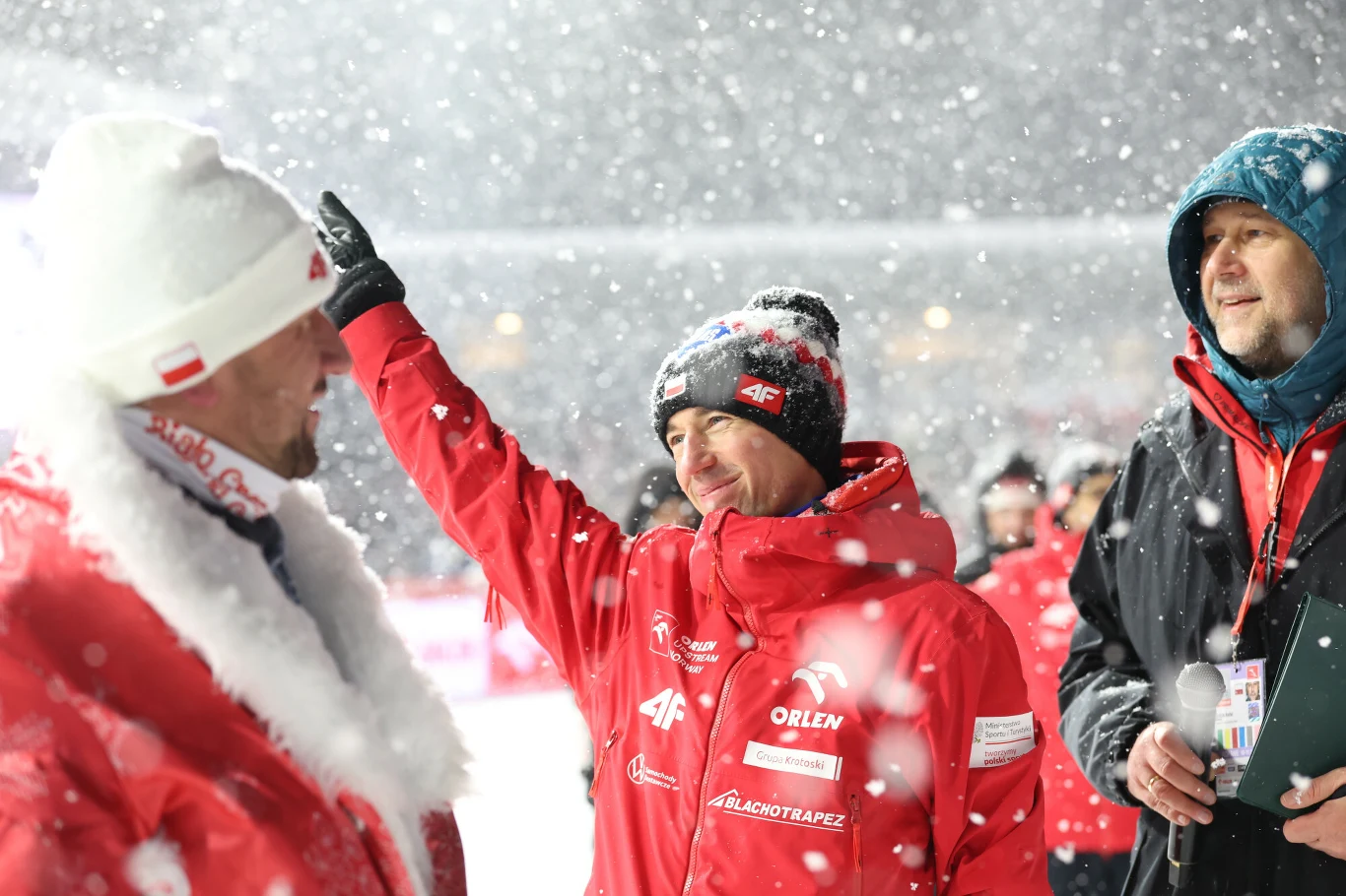 Kamil Stoch pożegnał się z kibicami Trzech mężczyzn ubranych w ciepłe kurtki i czapki podczas intensywnych opadów śniegu. Środkowy mężczyzna unosi rękę w geście pozdrowienia lub wskazywania, wszyscy wyglądają na zaangażowanych w rozmowę. W tle widać rozmyte sylwetki innych osób, całość r...
