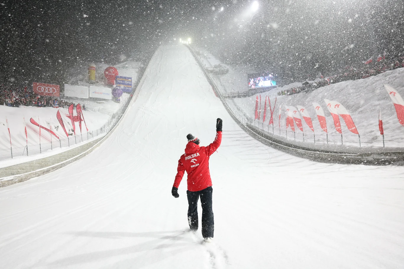 Kamil Stoch na Wielkiej Krokwi Osoba ubrana w czerwoną kurtkę idzie samotnie po zaśnieżonym torze skoczni narciarskiej nocą, podnosi rękę w górę, wokół pada gęsty śnieg, a z obu stron trasy stoją flagi i reklamy.