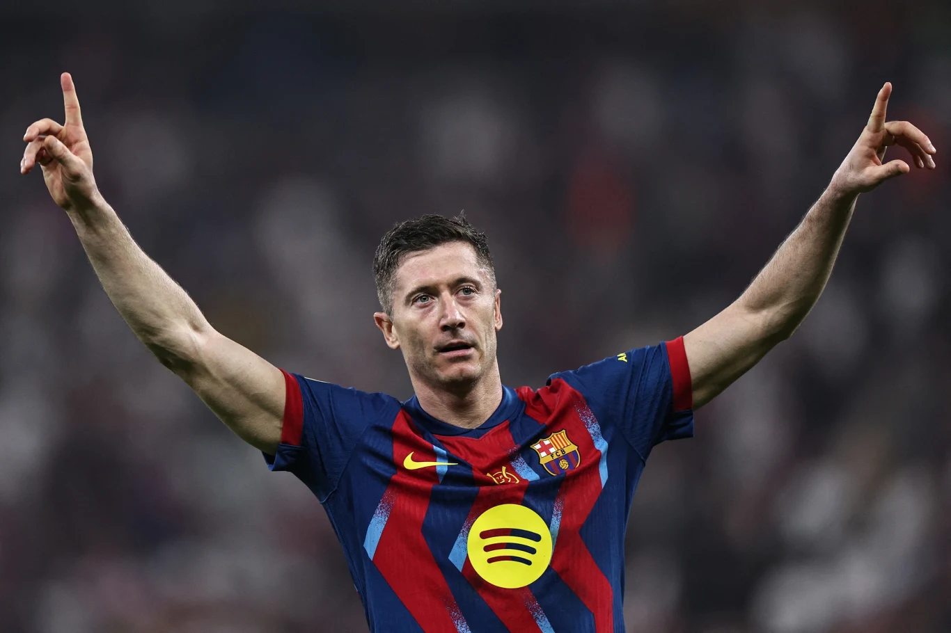 Piłkarz w stroju FC Barcelony z uniesionymi obiema rękami do góry, tło rozmyte, sugerujące stadion pełen kibiców.