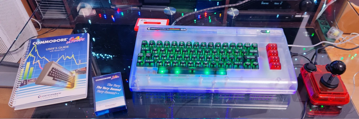 C64 Ultimate Stary komputer Commodore 64 z przezroczystą obudową i podświetlaną klawiaturą, joystick w stylu retro, instrukcja obsługi, kaseta magnetofonowa oraz dodatkowe akcesoria na szklanym biurku, w tle kolorowe lampki LED.