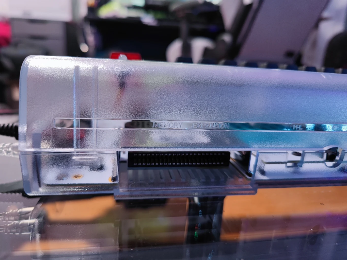 C64 Ultimate Przezroczysta obudowa z widocznym złączem elektronicznym, prawdopodobnie portem rozszerzeń lub pamięci, częściowo odbijająca światło z powierzchni pod spodem.