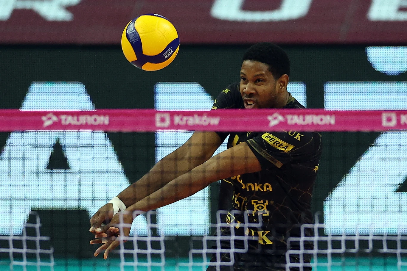 Zawodnik siatkarski w czarno-żółtym stroju odbijający piłkę podczas meczu, intensywnie skupiony na akcji, na pierwszym planie siatka oraz różowa taśma z napisem TAURON.