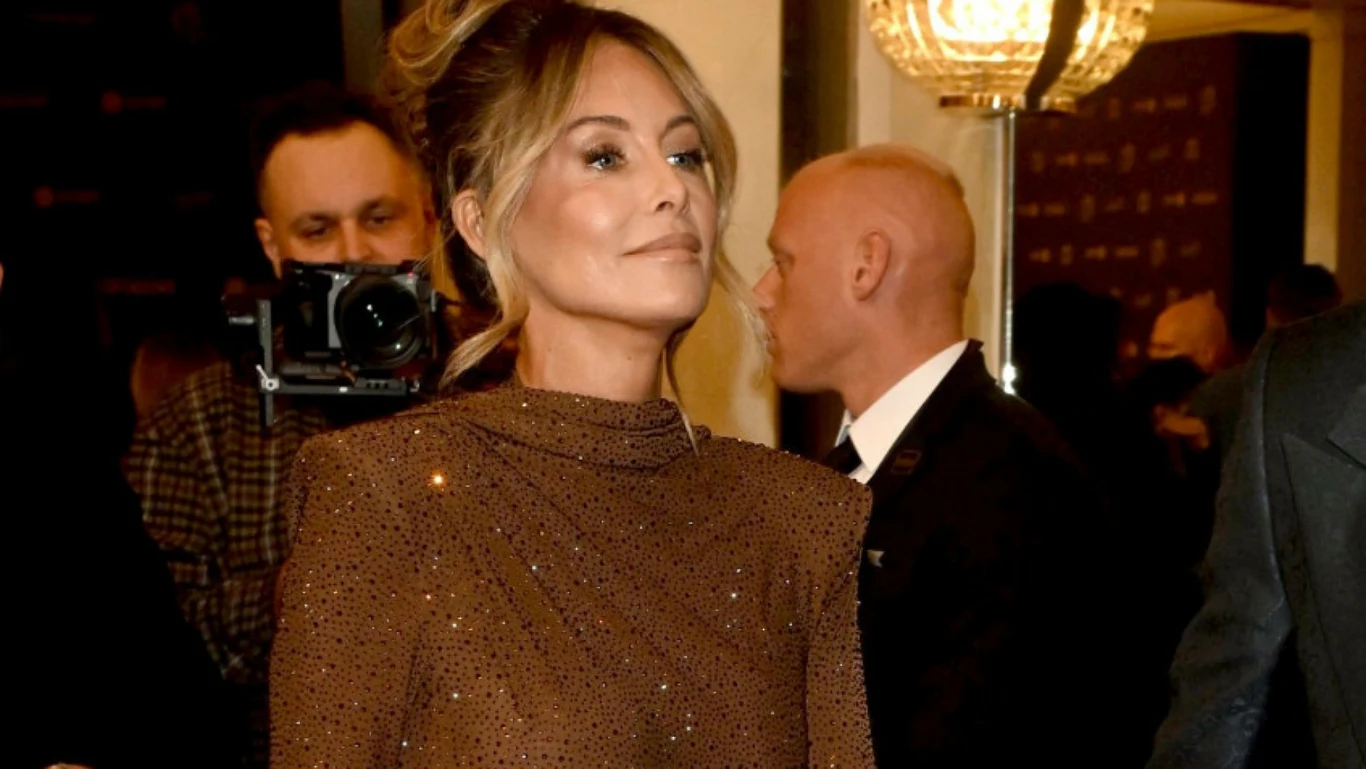 Kobieta w eleganckiej, błyszczącej sukni stoi w centrum uwagi podczas wydarzenia, w tle widoczni są inni uczestnicy oraz fotograf z aparatem, całość utrzymana w ciepłym oświetleniu.