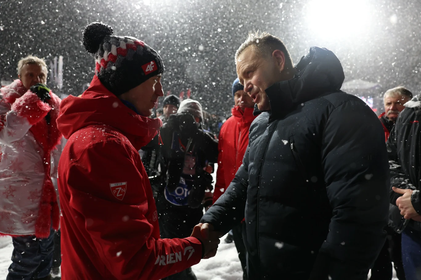 Dwóch mężczyzn w zimowej scenerii podaje sobie ręce, wokół nich stoi kilka osób w ciepłych ubraniach, z tyłu intensywnie pada śnieg i widać mocne światła.