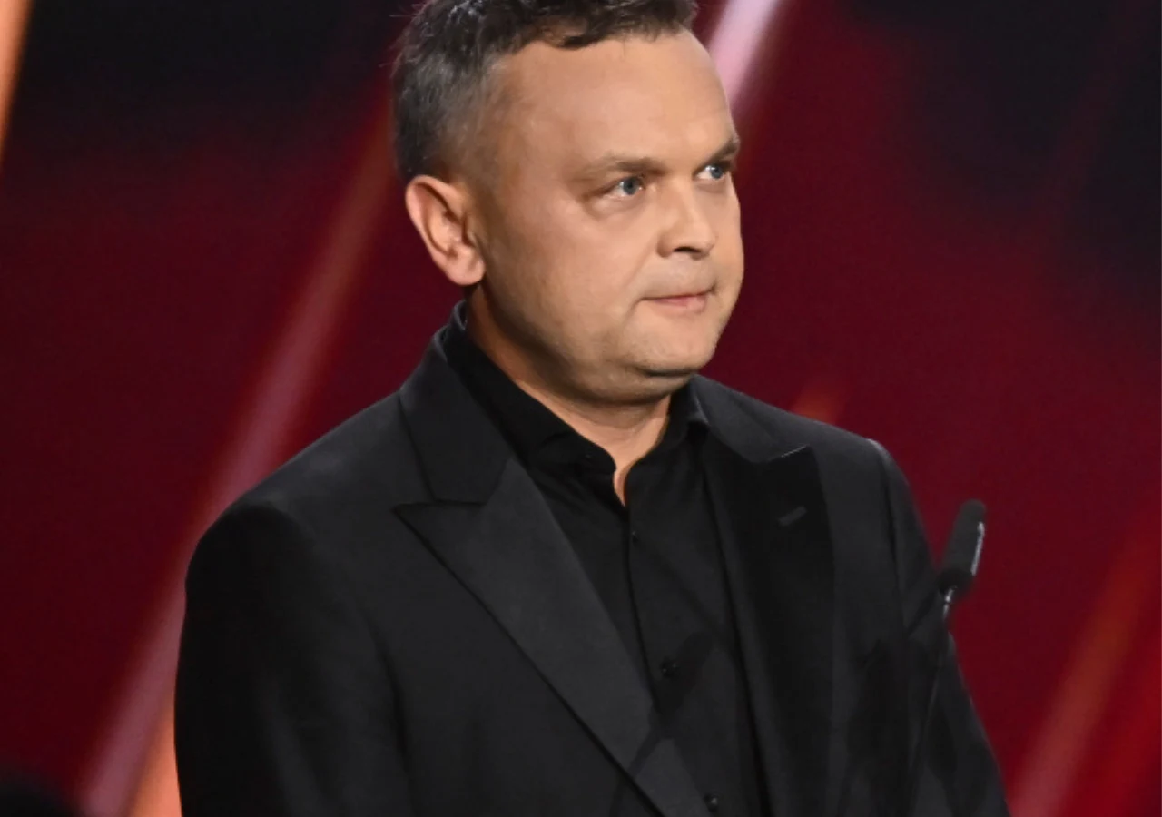 Tomasz Sygut - prezes TVP Mężczyzna w czarnym smokingu stoi przed mikrofonem na tle czerwonej scenografii z jasnymi, ukośnymi światłami.