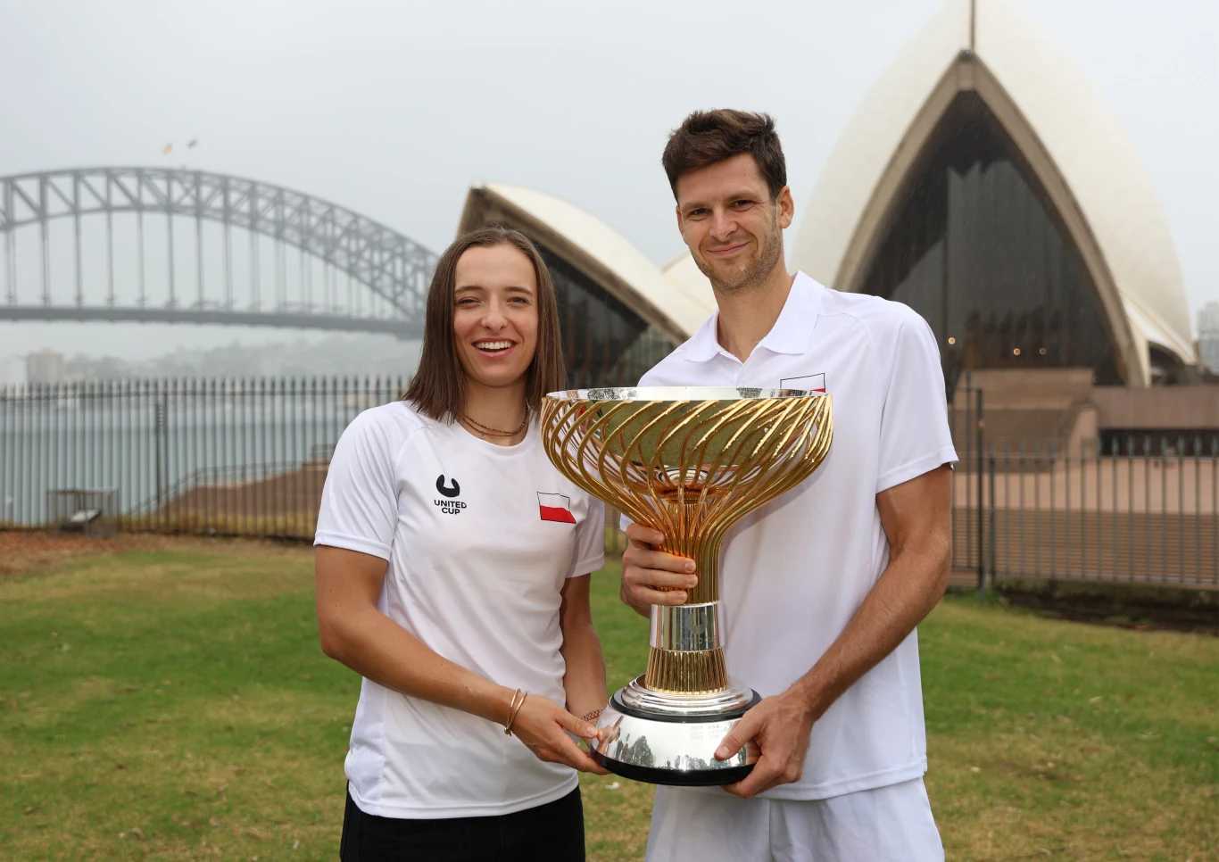 Dwójka sportowców w białych strojach trzyma dużych rozmiarów puchar. W tle charakterystyczne budynki Sydney Opera House i fragment mostu Sydney Harbour Bridge. Obie osoby uśmiechnięte, sceneria na zewnątrz.
