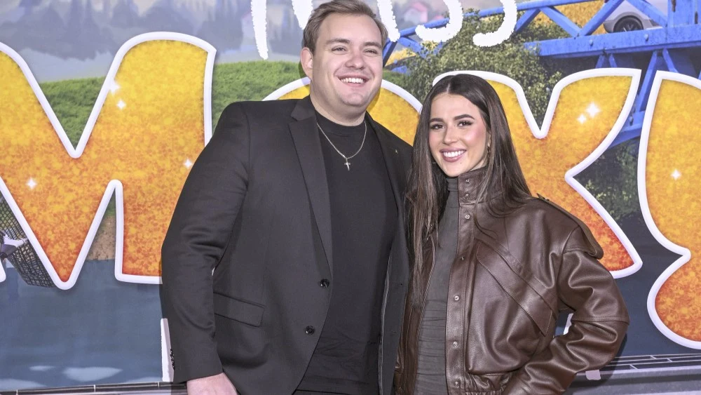 Roksana Węgiel i Kevin Mglej Mężczyzna ubrany w czarny garnitur i kobieta w brązowej skórzanej kurtce stoją razem, uśmiechają się do obiektywu na tle kolorowej, dekoracyjnej ścianki z dużymi literami.