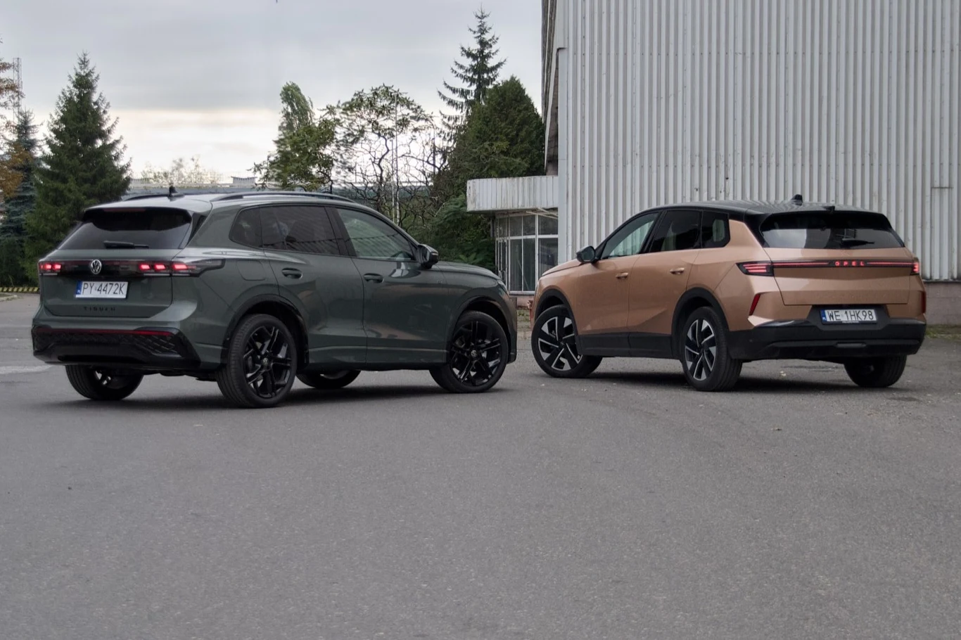 Dwa nowoczesne SUV-y stoją na otwartej przestrzeni asfaltowej obok przemysłowego budynku, w tle widoczne drzewa i lekko zachmurzone niebo.