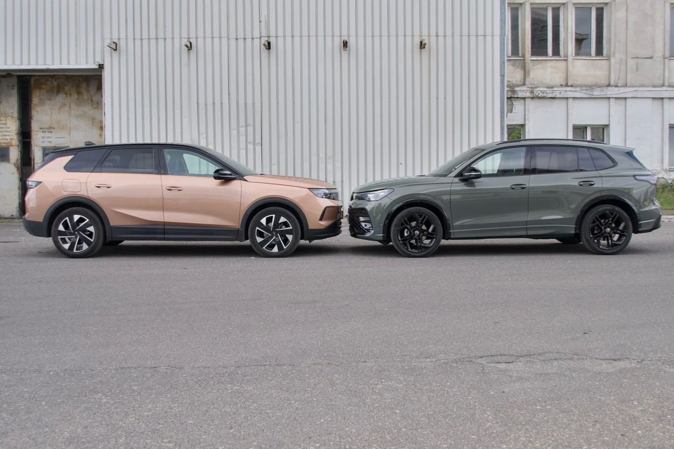 Dwa nowoczesne samochody SUV ustawione przodem do siebie na tle szarego przemysłowego budynku. Kolory pojazdów to matowy różowy oraz metaliczny szary, oba auta mają ciemne szyby i charakterystyczne felgi.