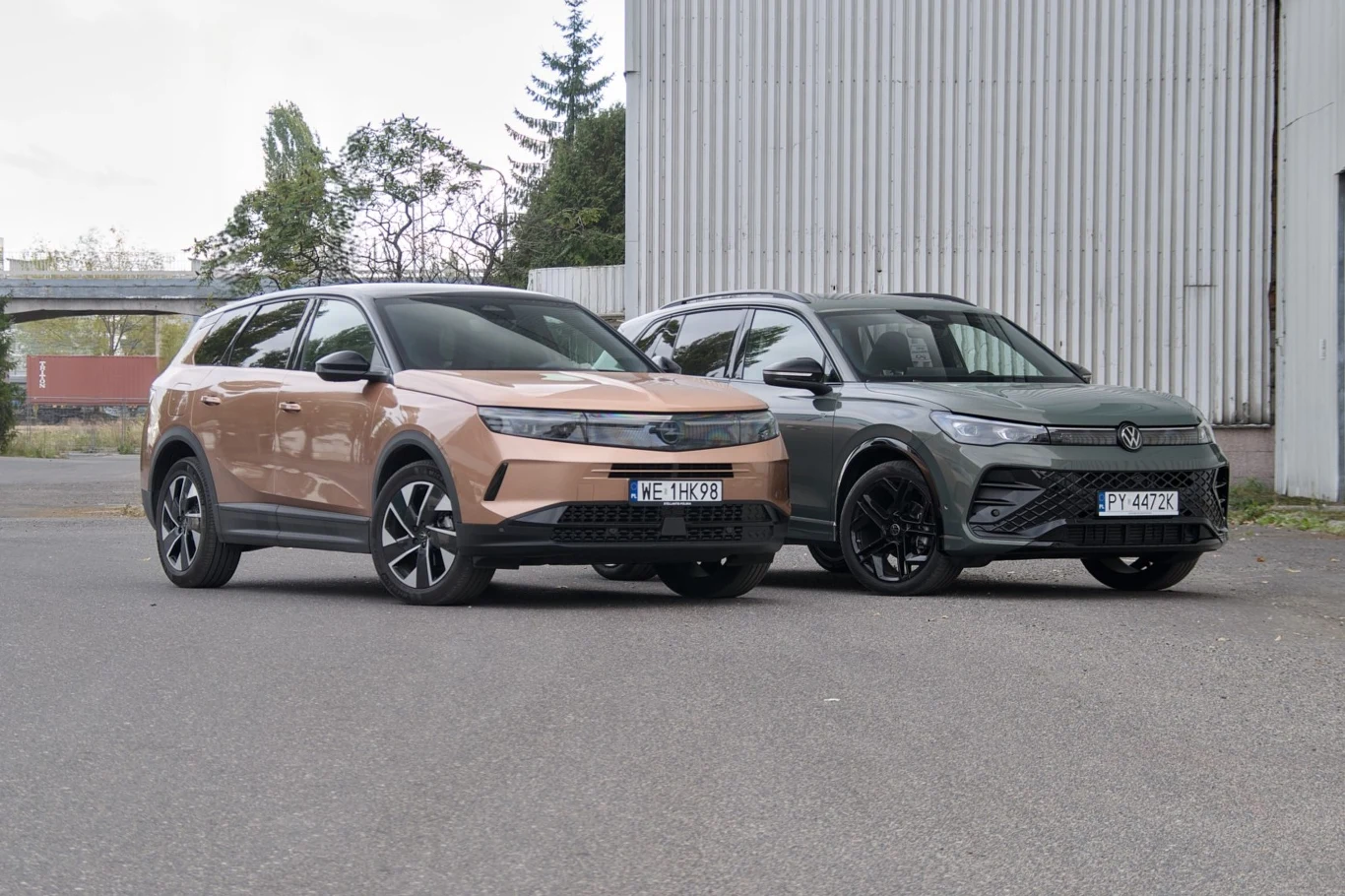 Dwa nowoczesne SUV-y zaparkowane obok siebie na tle metalowej, przemysłowej ściany. Pierwszy samochód jest jasnobrązowy, drugi ciemnoszary. Oba pojazdy ustawione są przodem do obserwatora.