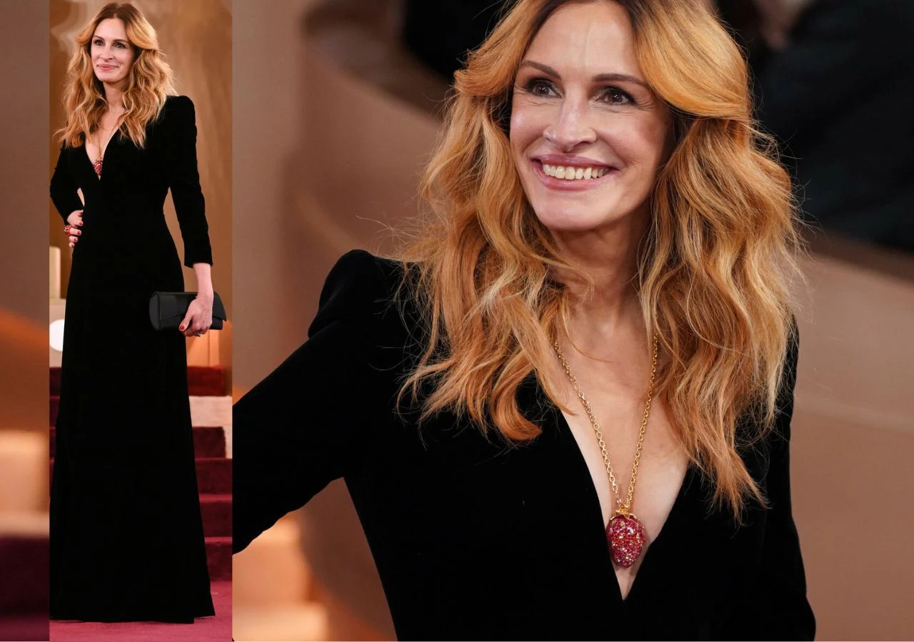 Kobieta o jasnych, falowanych włosach ubrana w długą, czarną suknię z głębokim dekoltem trzyma czarną kopertówkę i uśmiecha się, pozując na tle schodów. Szyję zdobi długi naszyjnik z dużym, czerwonym wisiorem.