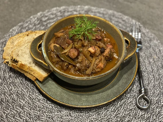 Gorący bigos z kawałkami mięsa i kiełbasy, podany w ceramicznej misce, udekorowany świeżym koperkiem, obok plastry chleba i widelec na teksturowanej podkładce.