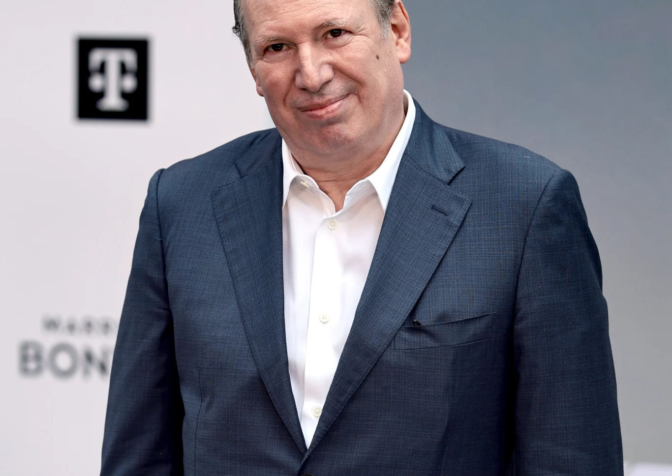 Hans Zimmer odpowiedział na skróconą transmisję Złotych Globów Mężczyzna w średnim wieku w granatowej marynarce i białej koszuli, stojący na tle jasnej ścianki z widocznymi logotypami.