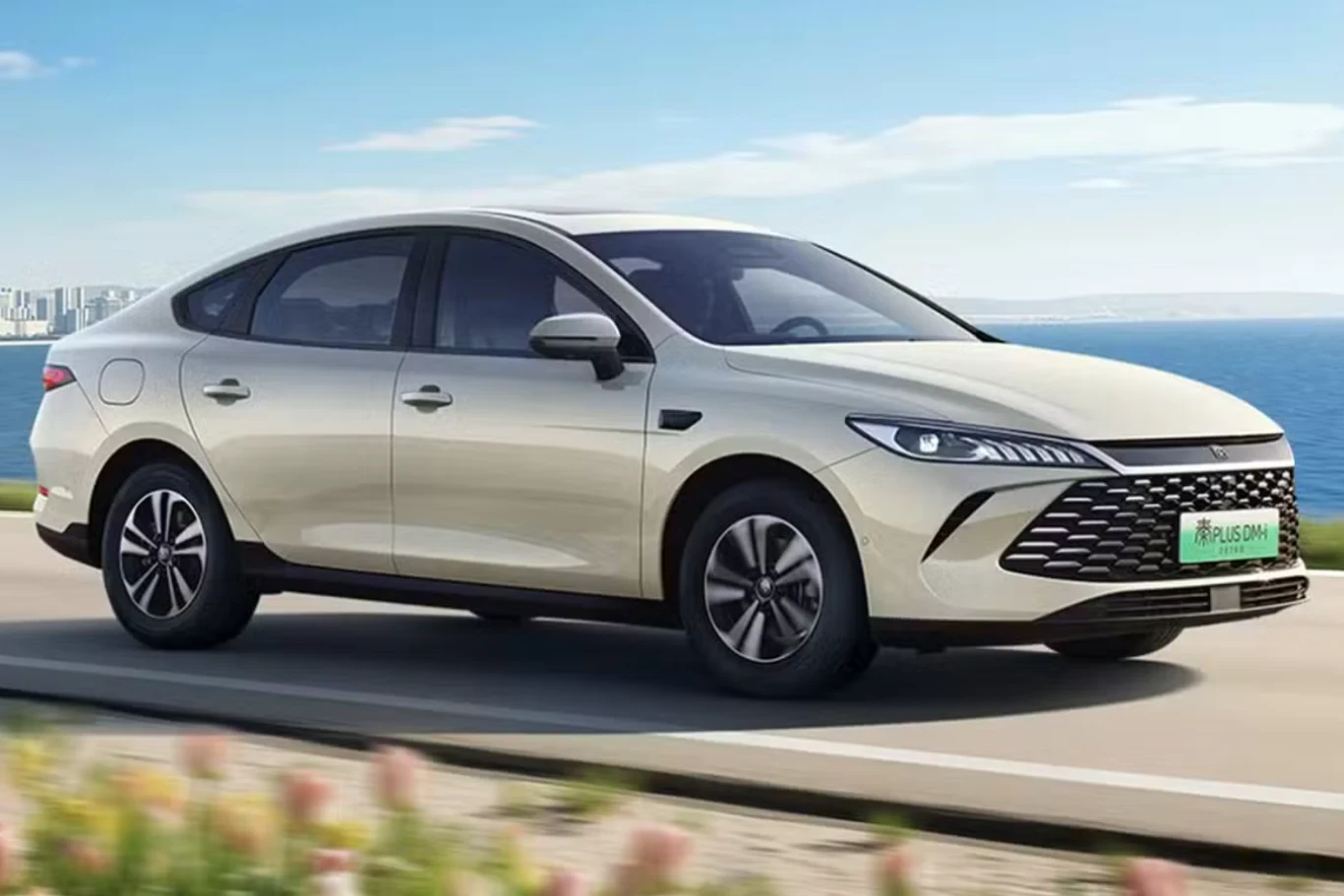 Jasny nowoczesny sedan poruszający się po drodze w słoneczny dzień, w tle widoczny fragment wybrzeża z morzem i niebem, dookoła roślinność, a auto wyróżnia się dynamiczną sylwetką i wyrazistym przednim grillem.