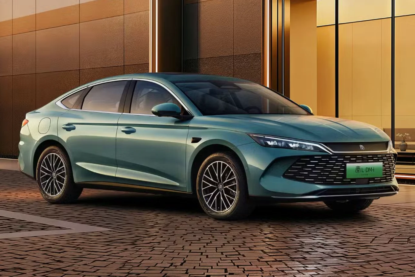 Nowoczesny samochód osobowy w kolorze błękitno–zielonym stoi zaparkowany na brukowanej nawierzchni na tle budynku z przeszklonymi ścianami. Auto wyróżnia się dynamiczną sylwetką, designerskimi reflektorami LED oraz eleganckimi felgami.