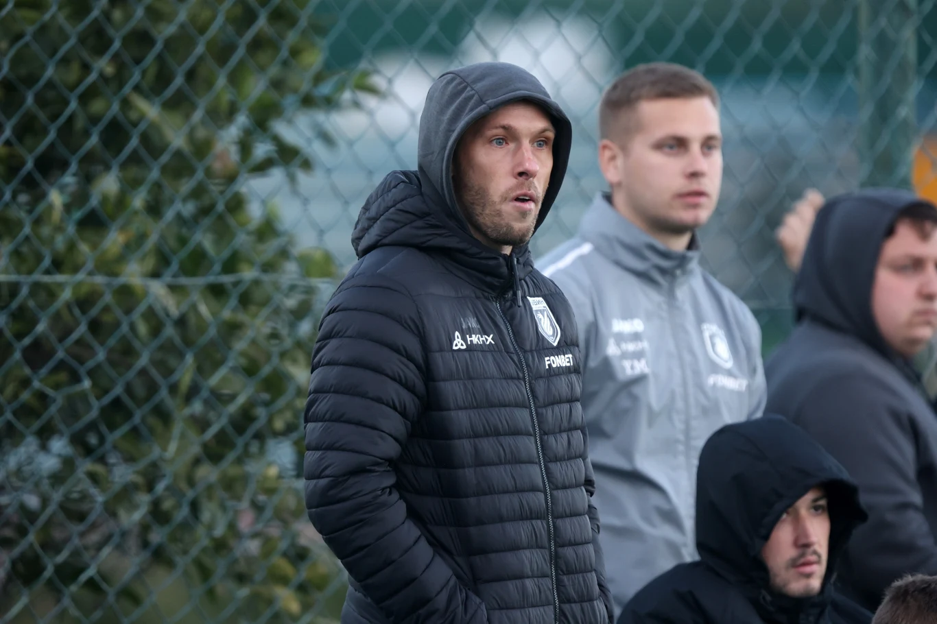 Grupa młodych mężczyzn ubranych w sportowe kurtki oraz bluzy stoi na zewnątrz przy siatce ogrodzeniowej, jeden z nich ma kaptur na głowie i trzyma ręce w kieszeniach.