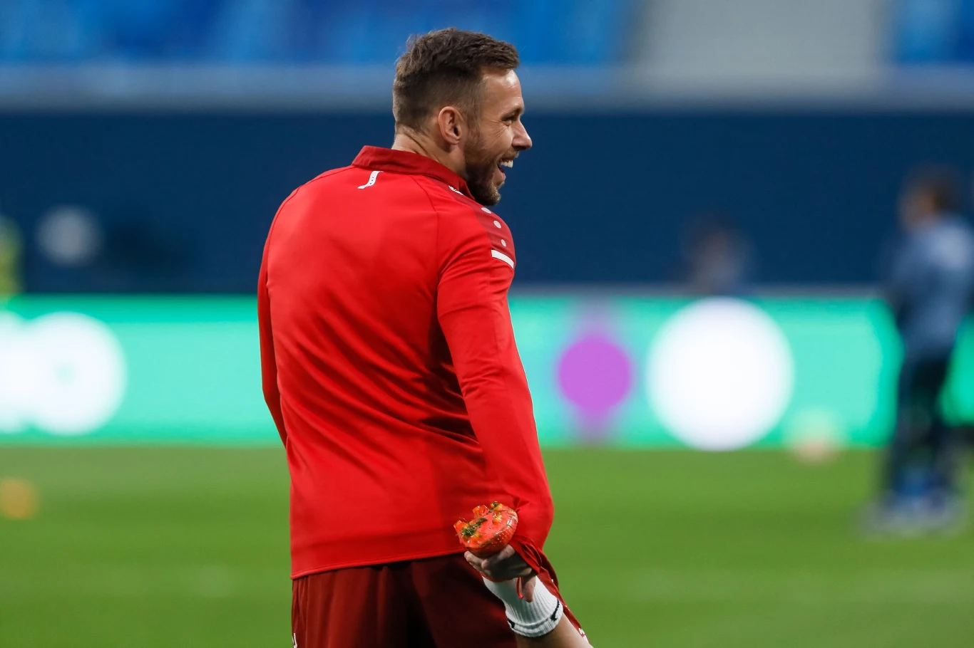 Mężczyzna w stroju sportowym stoi na boisku piłkarskim, uśmiechając się i trzymając rękawice bramkarskie. W tle zamazane kontury stadionu oraz innych osób.